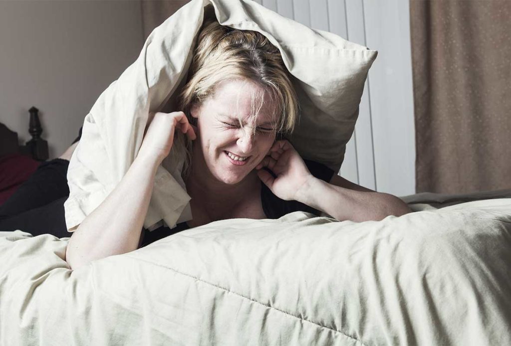 A woman sitting up in bed with a pillow on her head and ger fingers blocking her ears.
