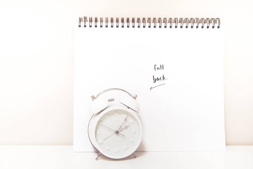 A white clock in front of a notepad that has 'fall back' written on it.