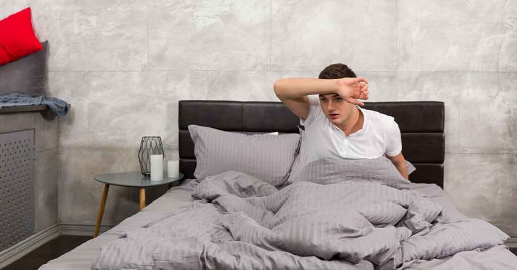 A man sitting up in bed wiping sweat off his forehead