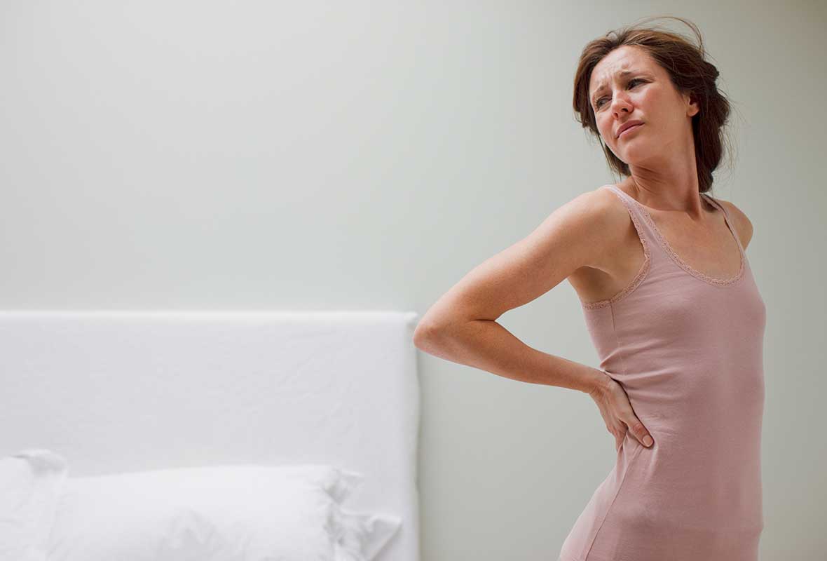 A young woman standing next to a bed wincing with back pain