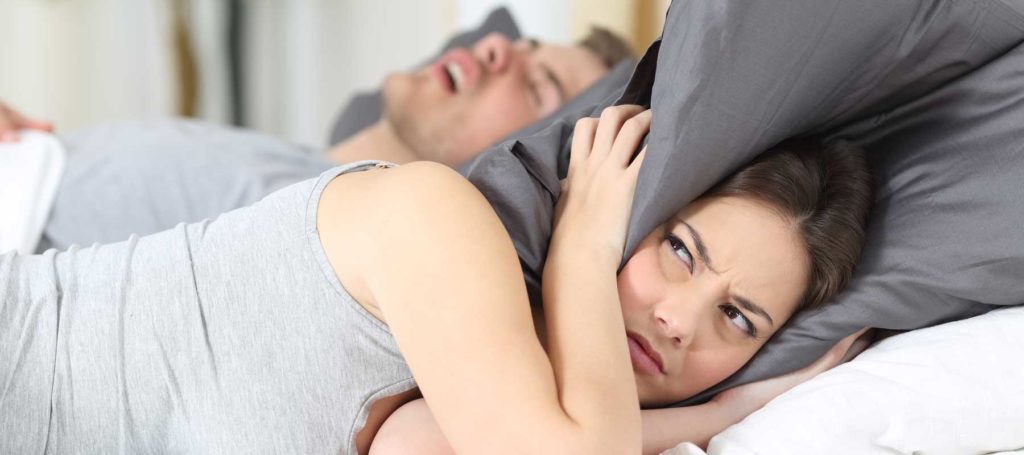 A man and woman lying in bed. The woman has a pillow wrapped around her head while the man snores.