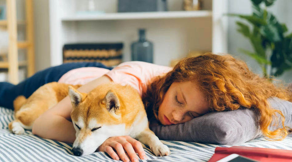 Woman lying on a bed, her head on a pillow and a dog lying on her arm.