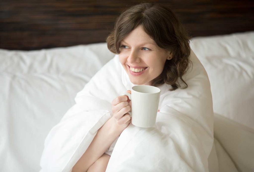A woman sitting on her bed, snuggled up in a blanket, holding a mug