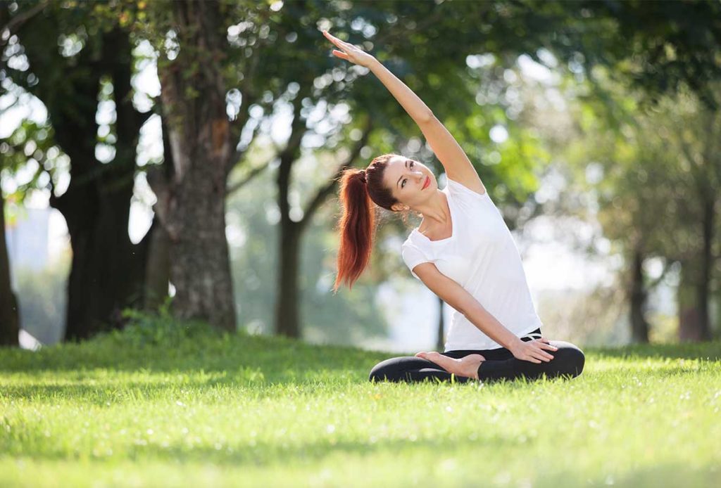 A woman sitting in the park near trees, doing yoga stretches.