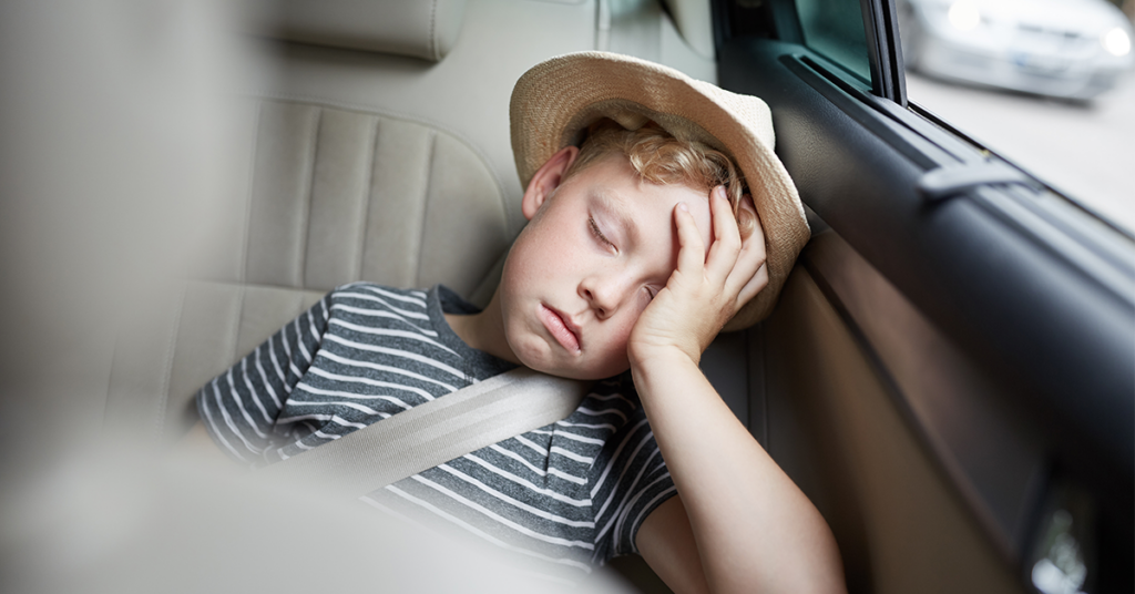 A young boy wearing a hat, sleeping in a car with his head in his hand