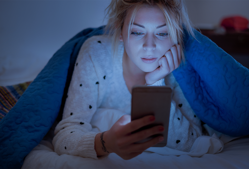 A tired woman under a blanket looking at her phone screen in bed