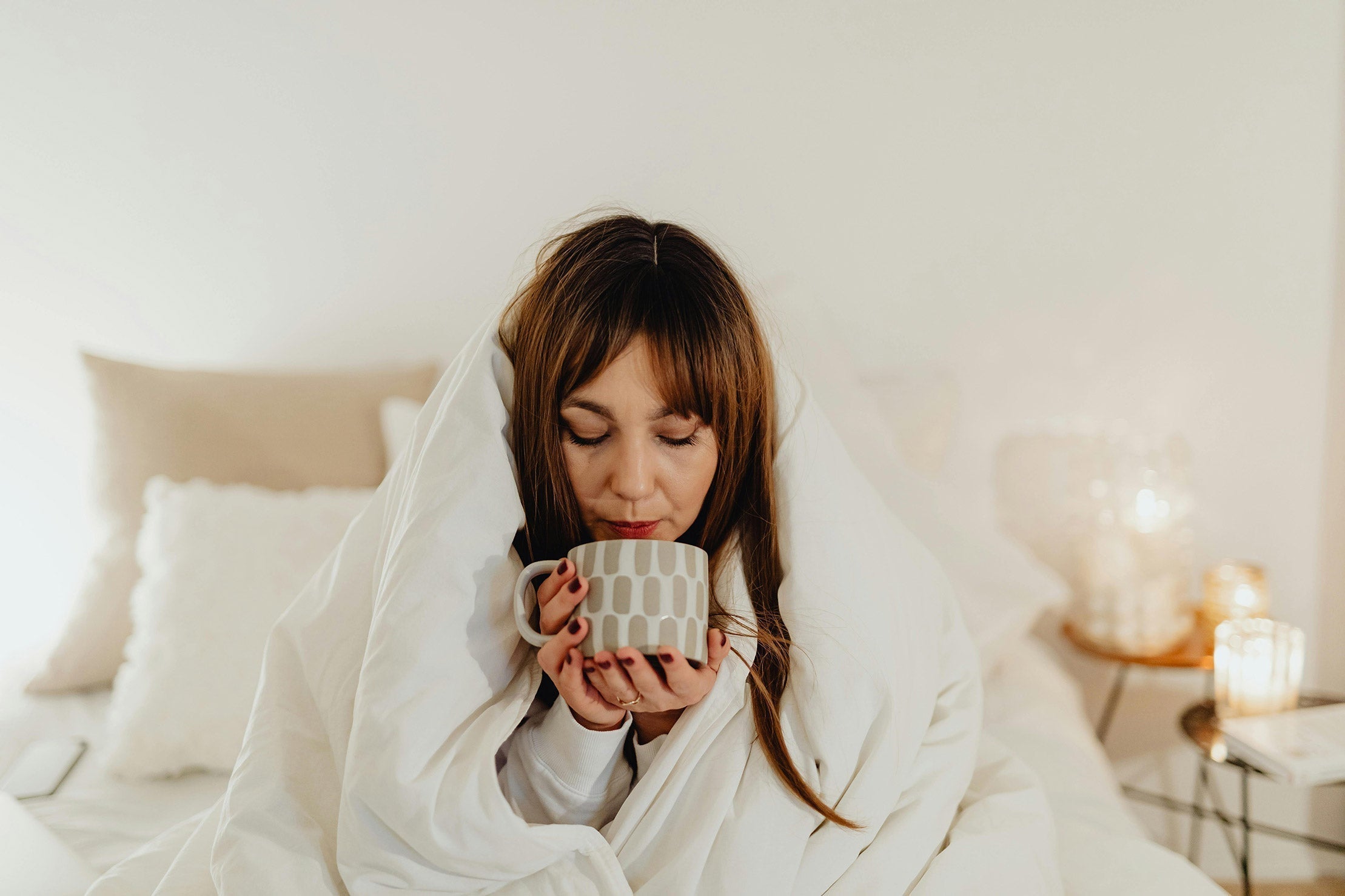 A woman sitting in bed, covered in a blanket, drinking a mug of tea