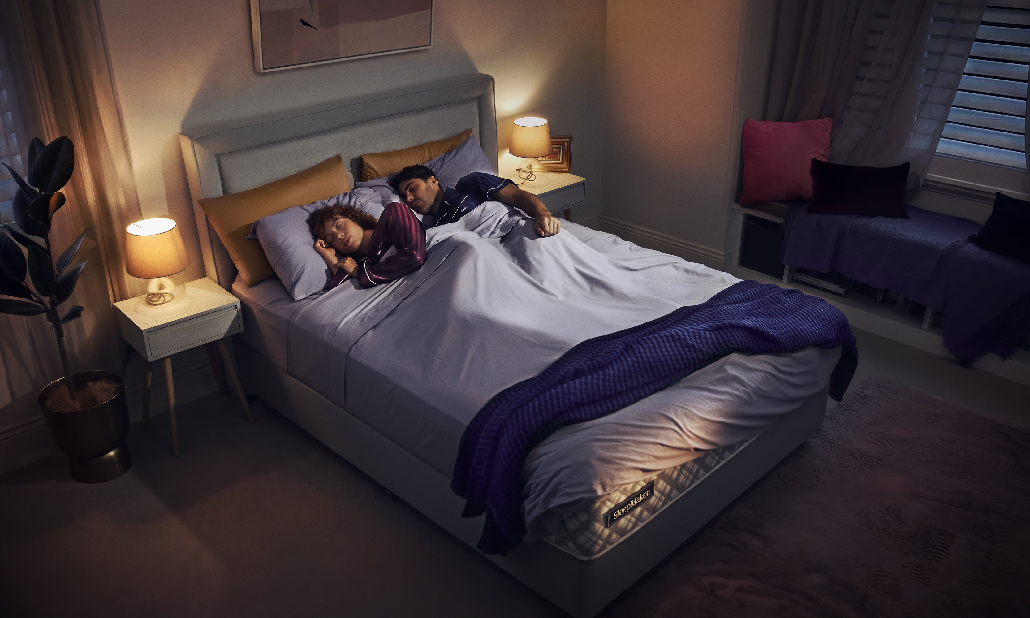 Two people lying in a SleepMaker bed under a blanket in a dimly lit bedroom.