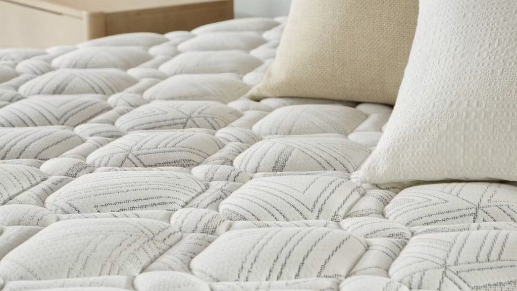 A close-up of a SleepMaker quilted mattress, styled with two cream pillows in a softly lit bedroom setting.