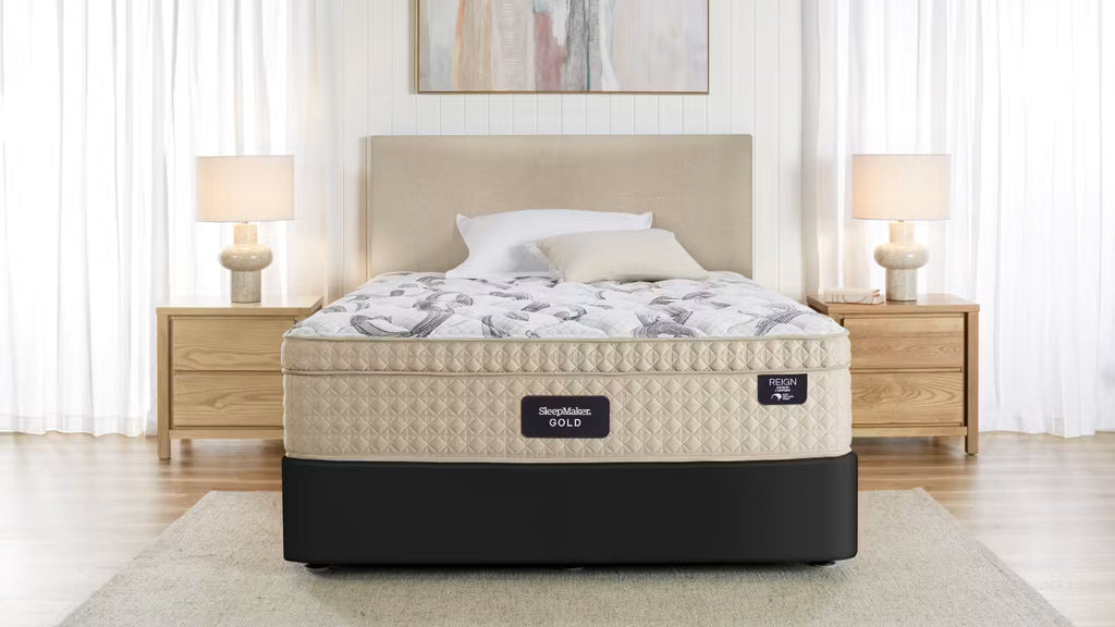A bed with a SleepMaker Gold mattress, styled in a bright, modern bedroom with wooden nightstands, lamps, and a neutral beige headboard.