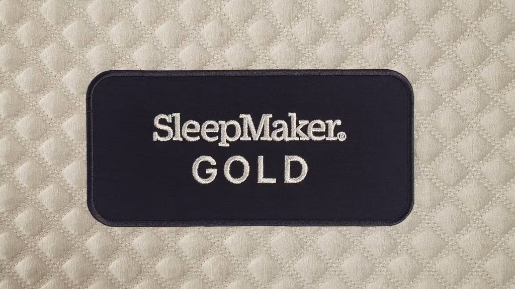 A close-up of a mattress with a quilted diamond pattern in a light warm neutral tone, featuring a dark navy rectangular label with the words 'SleepMaker GOLD' in white text.