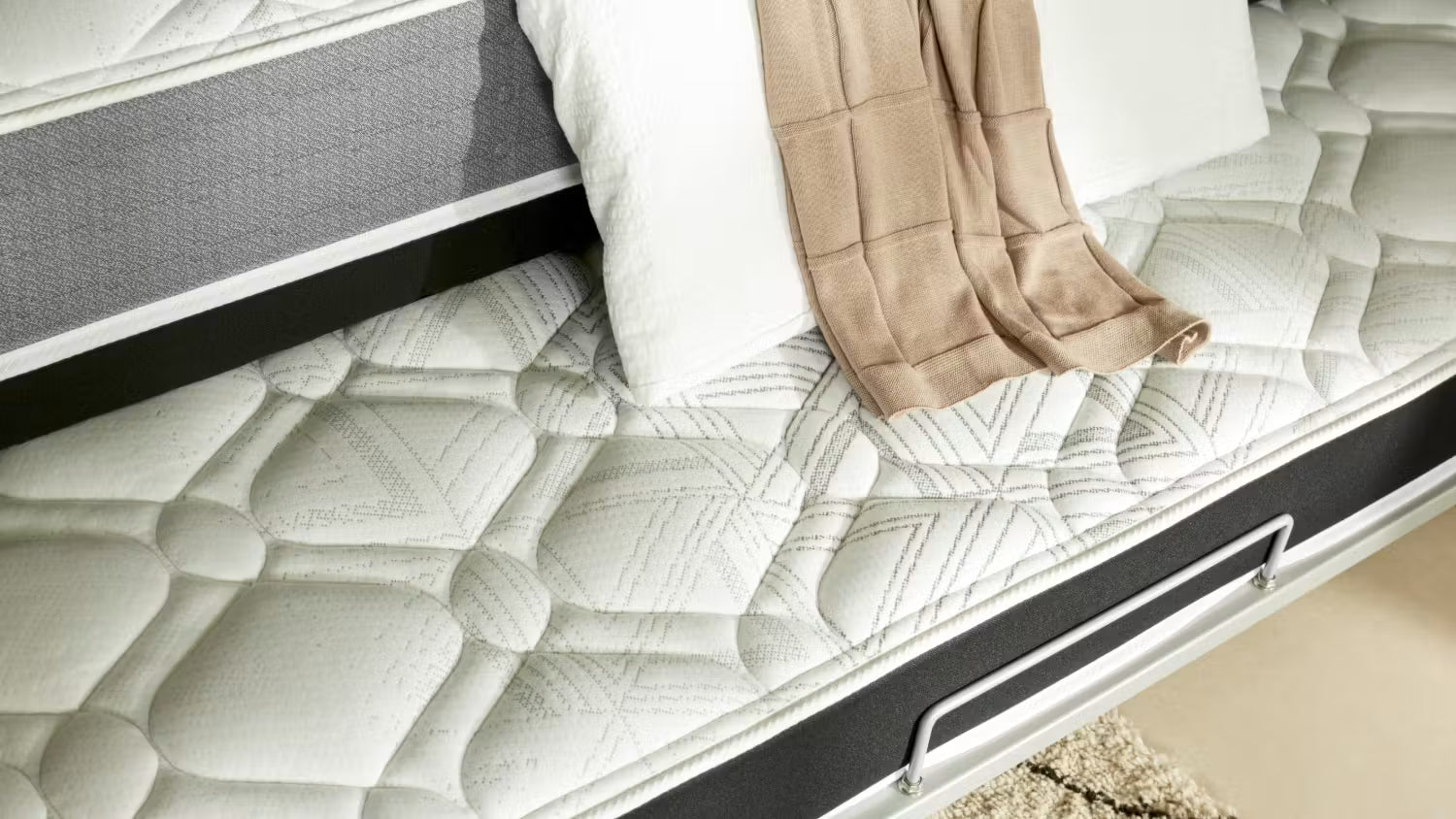Close-up of a SleepMaker trundler bed in off-white with a geometric diamond pattern, shown on a bed frame with white pillows and a beige throw blanket.