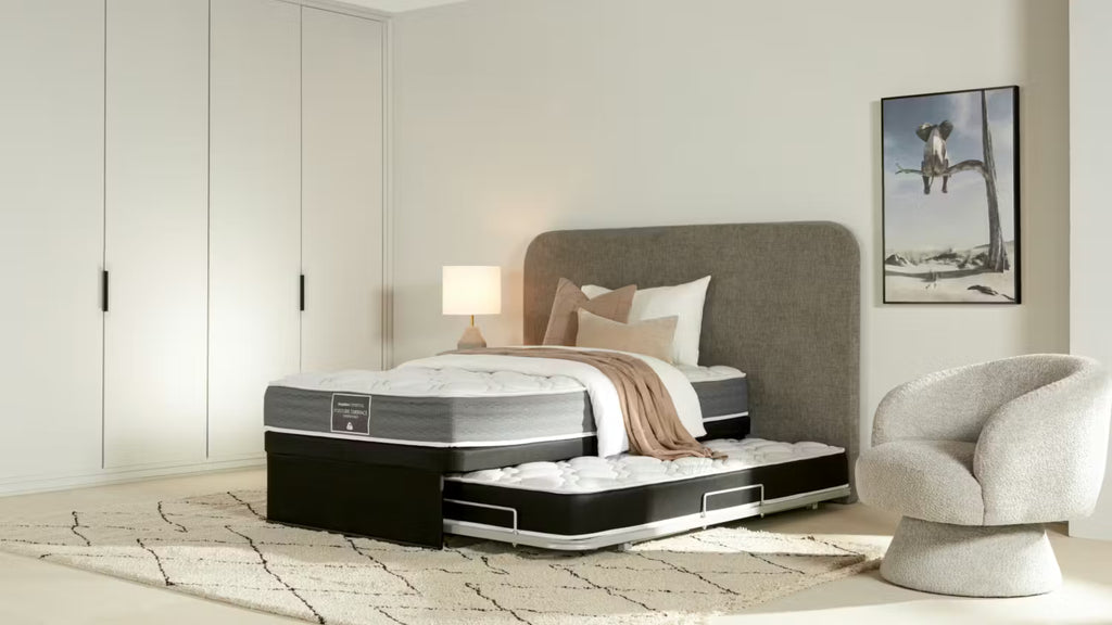 A SleepMaker trundler bed with a gray upholstered headboard and a matching under-bed trundle, styled in a modern bedroom with a gray armchair, white wardrobe, and framed wall art.