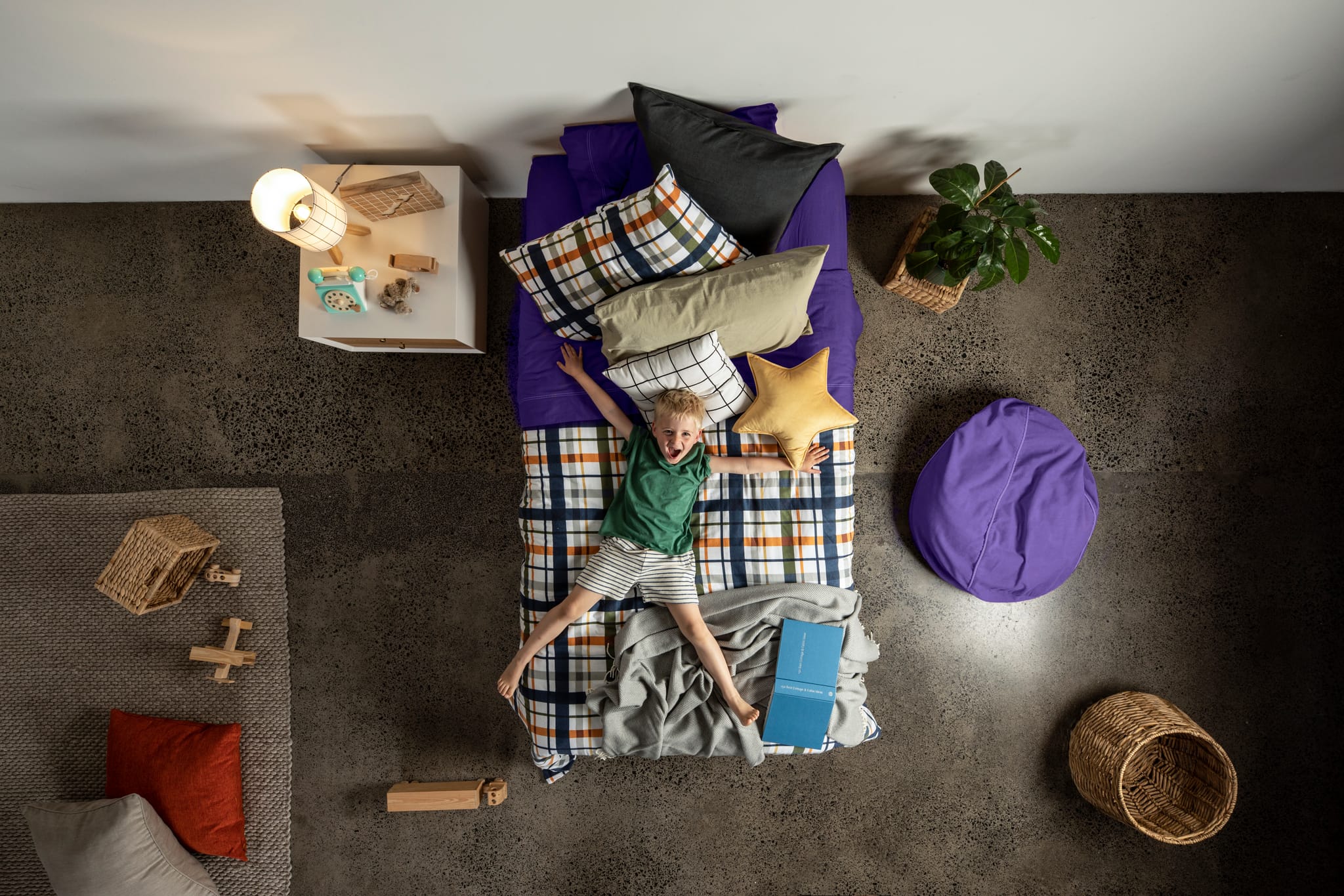Child playing on a colorful SleepMaker bed with pillows and blankets in a room with a rug and small table.