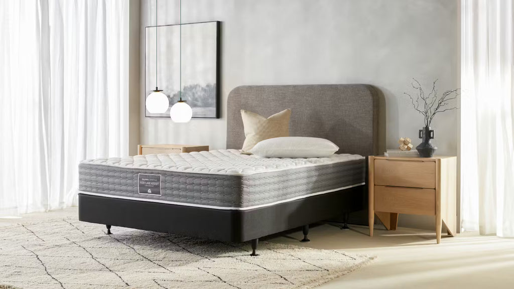 A SleepMaker Posture Assist bed with a gray upholstered headboard and a quilted gray-and-white mattress on a dark base, styled in a bright bedroom with a wooden nightstand, curtains, and a rug.